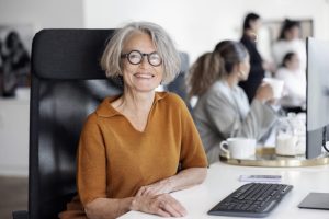 lächelnde ältere Frau mit Brille, grauen Haaren und einer Beigen Bluse sitzt an einem Schreibtisch in einem Meeting-Raum.