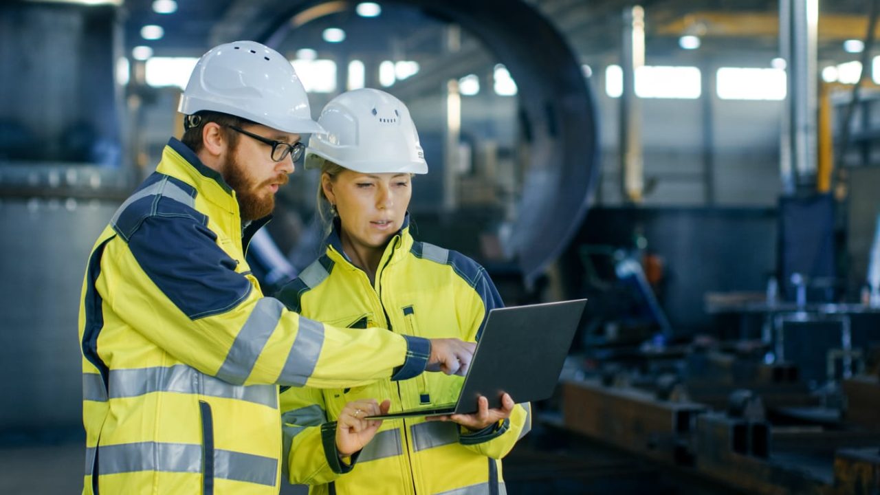 Ein Mann und eine Frau mit weißem Helm und grüner Warnweste schauen auf einen Laptop