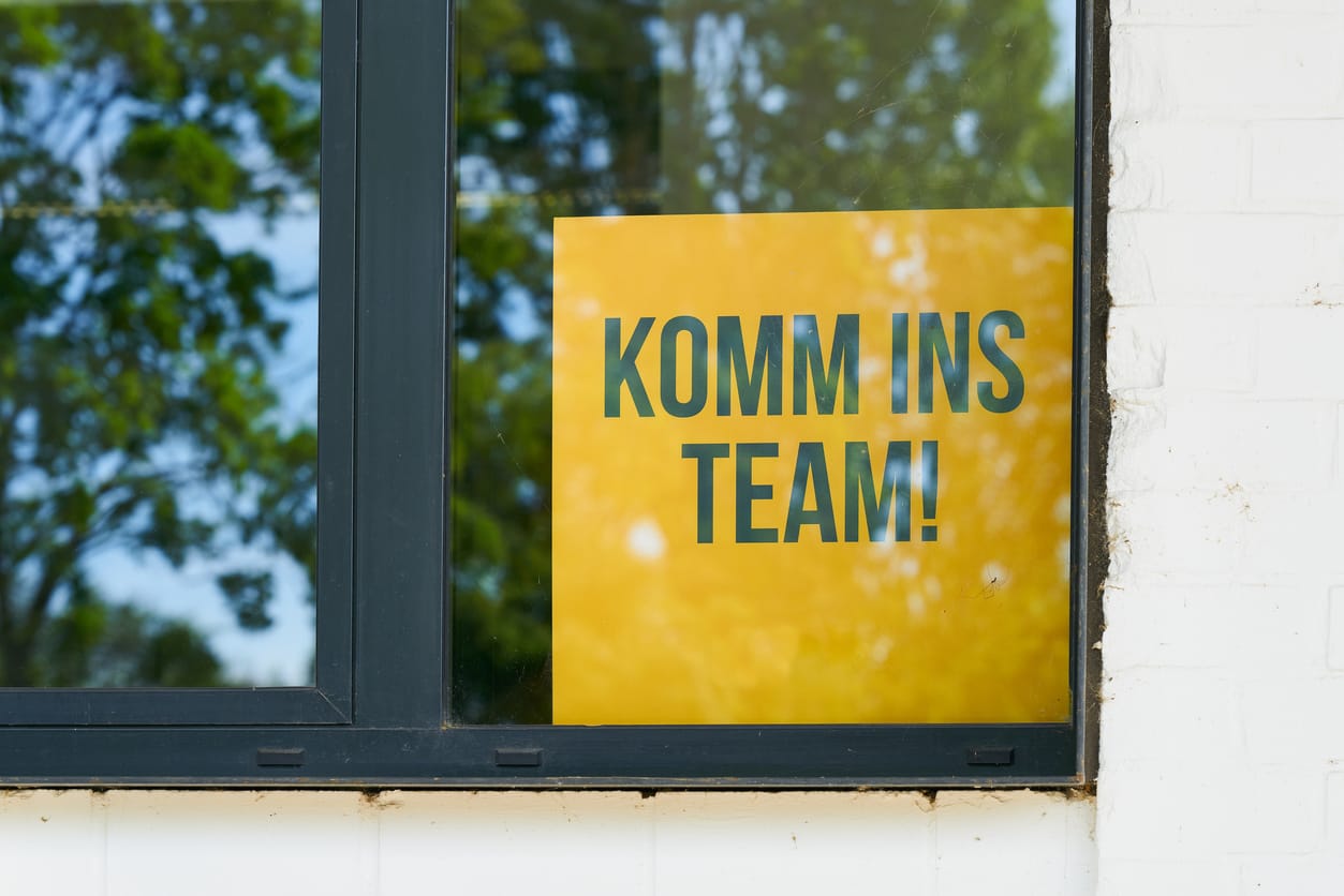 Gelbes Plakat mit der Aufschrift ‘KOMM INS TEAM!’ im Fenster, umgeben von Bäumen in der Spiegelung. Steht sinnbildlich für modernes Recruiting, aktive Personalsuche und eine einladende Teamkultur.