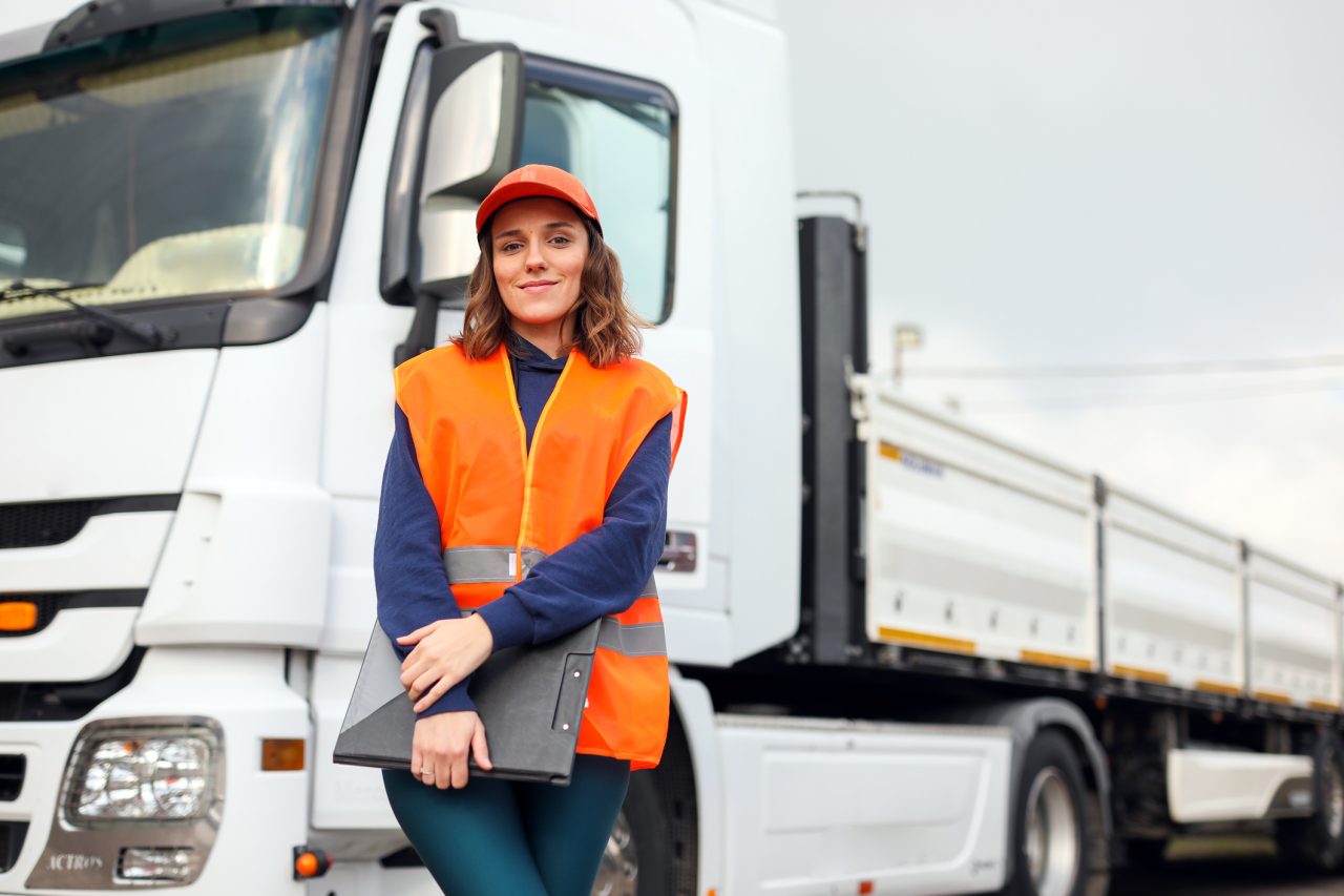 Junge Frau mit langen braunen Haren, orangener Mütze, orangener Warnweste und Klemmbrett in der Hand steht vor einem weißen LKW