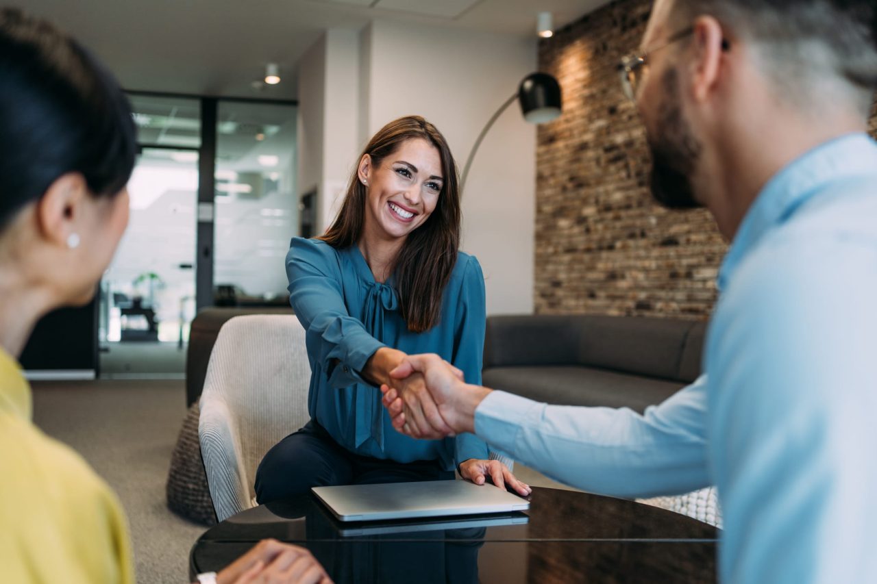 Lächelnde Bewerberin in einer modernen Büroumgebung, die bei einem Vorstellungsgespräch einem potenziellen Arbeitgeber die Hand schüttelt und so beruflichen Erfolg, Professionalität sowie eine positive Arbeitsatmosphäre symbolisiert.