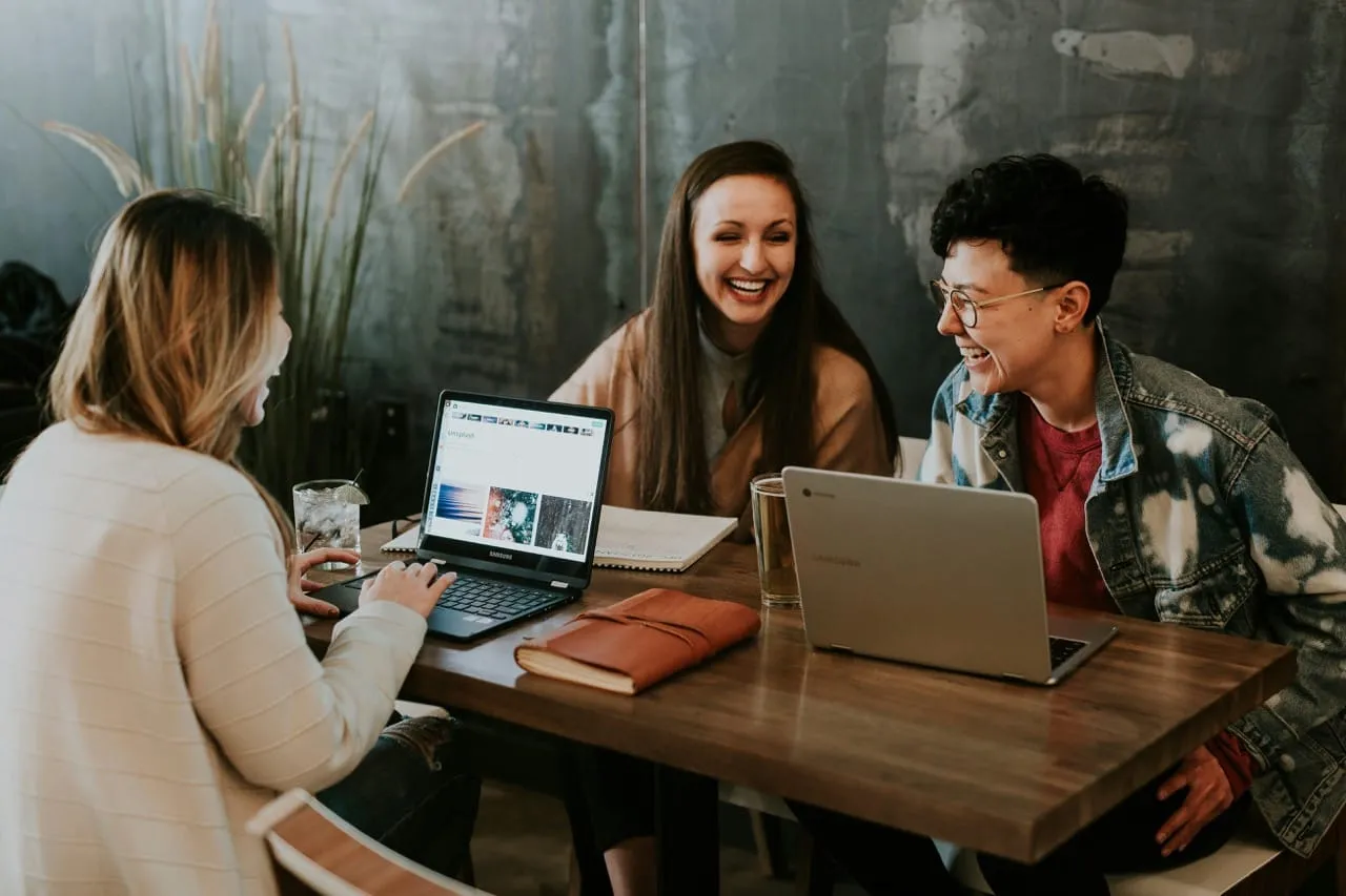 Drei junge Berufstätige in entspannter Atmosphäre lachen und arbeiten gemeinsam an Laptops in einem modernen Café.