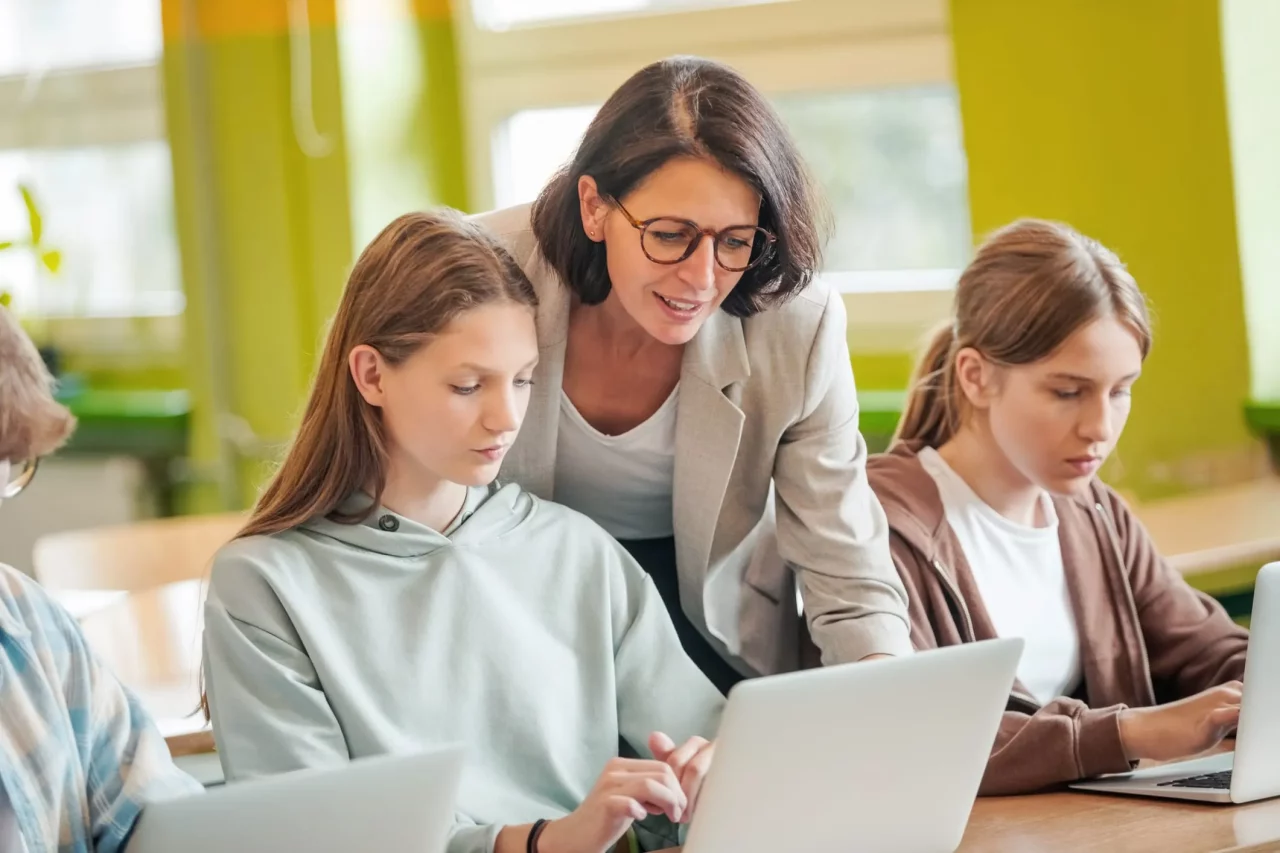 Lehrerin unterstützt Schülerinnen beim Arbeiten am Laptop in einem modernen Klassenzimmer mit grünen Wänden.